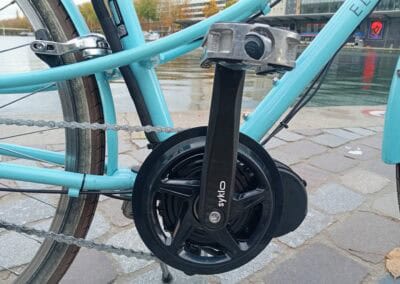 Vélo électrique bleu, moteur SYKLO, à proximité d'une rivière et d'un bâtiment moderne.
