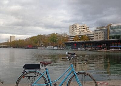 Vélo électrique bleu au bord de la Seine, Paris, avec bâtiments modernes et ciel nuageux.